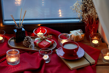 holidays, decoration and celebration concept - christmas gift, cup of coffee, candles and aroma reed diffuser on red tablecloth on window sill at home