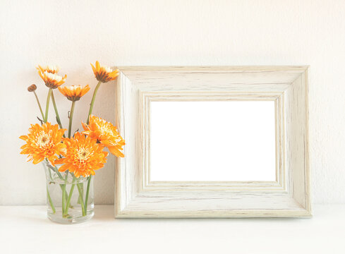White wooden rectangle photo frame and orange flower (chrysanthemum) in vase on a wooden shelf with a white wall as background...