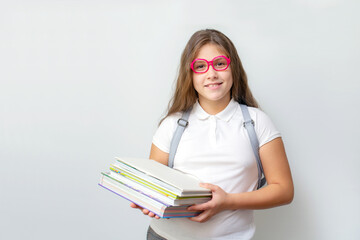 Portrait of smiling little schoolgirl in pink glasses with backpack holding stack of books on white background