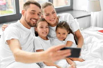 family and people concept - happy mother, father and two daughters taking selfie with smartphone in bed at home