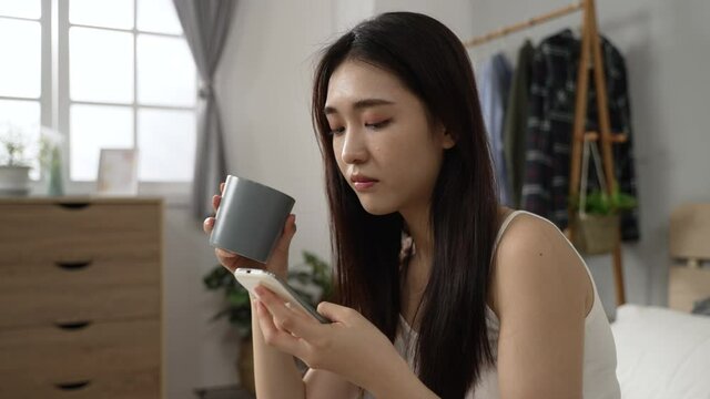 confused Korean woman staring at phone screen and checking the ridiculous posts on social media with a funny face while having coffee in the morning at home