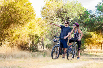Two cyclists, a man and a woman, standing with their bicycles in the middle of the countryside. The male cyclist points with his finger to the female cyclist the place where they should arrive