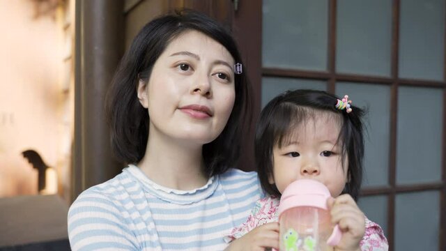 closeup shot loving mother looking at the toddler girl in arms and into distance as she is drinking water from sipper cup at their traditional japanese house