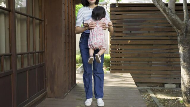back view with slow motion japanese toddler girl in kimono is waddling along outdoor corridor of her house towards smiling mother who bends down to lift her up.