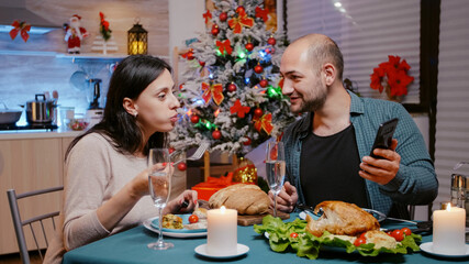 Man and woman eating festive dinner looking at smartphone for entertainment on christmas eve. Couple with gadget enjoying meal and glasses of champagne. People using technology on holiday