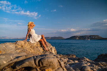 Beautiful young woman wearing traw hat sitting in rocks in white dress by the seashore