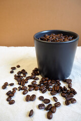 on a beige background of a table with scattered coffee beans, there is a dark mug filled with roasted whole coffee beans. side view