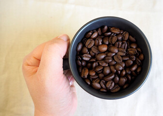 a woman in a beige wool sweater holds a dark gray mug with whole coffee beans on a brown background. top view. copy space
