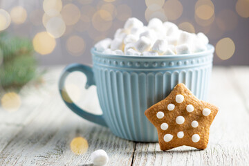 Fototapeta na wymiar A blue cup of winter hot drink with marshmallows and gingerbread star on a white wooden background with bokeh.