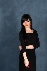 Fototapeta premium Expressive and glamorous beautiful girl, brunette in a black dress. Beautiful and slender woman sad and thoughtful, studio portrait on a monochrome blue, trendy shabby background