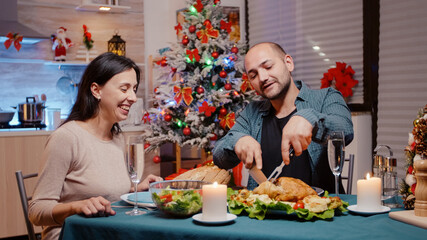 Man cutting chicken for woman at festive dinner on christmas eve festivity. Cheerful couple enjoying traditional meal and drinking champagne from glasses on table. Seasonal celebration
