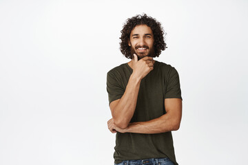 Portrait of thoughtful handsome middle-eastern man, smiling and looking pleased, thinking of smth interesting, standing over white background