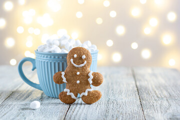 A blue cup of winter hot drink with marshmallows and gingerbread man on a white wooden background with bokeh.