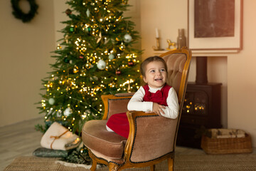 Obraz na płótnie Canvas A cute cheerful laughing child is sitting in an armchair by the fireplace against the background of a Christmas tree with lights. Cozy room with Christmas decorations