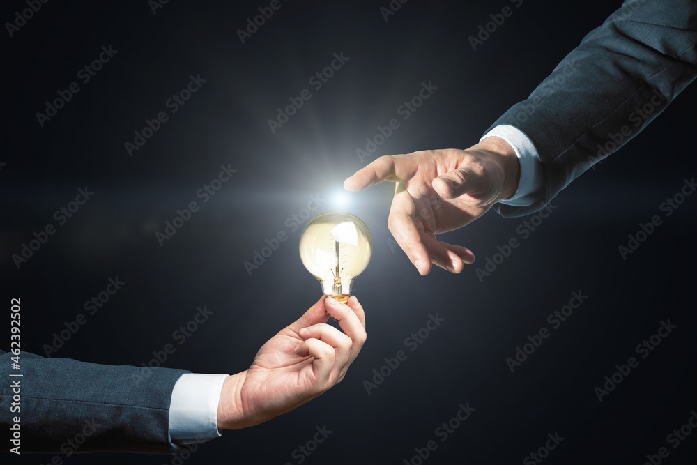 Poster close up of two businessmen hands pointing and and holding lamp on black background. innovation, ide