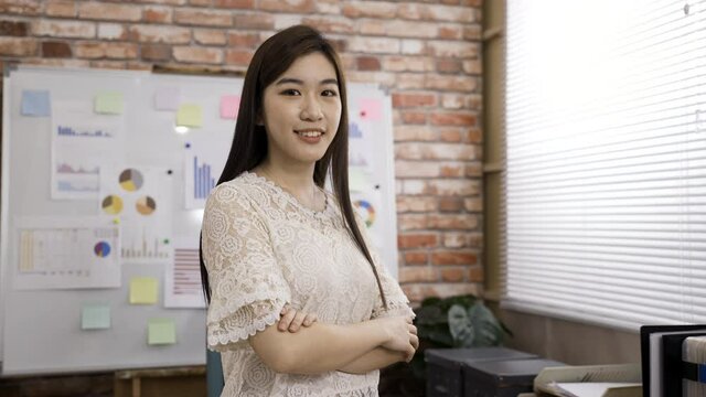 pretty confident asian female manager is smiling at the camera with crossed arms in a bright loft office at daytime.
