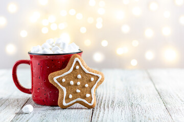 Obraz na płótnie Canvas A red cup of winter hot drink with marshmallows and gingerbread star on a white wooden background with bokeh.