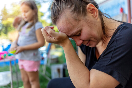 Stressed depressed young mother sits outside in a playground with her kids playing behind her. Single frustrated woman hold her head with hands sitting on chair with playful kids on a background.
