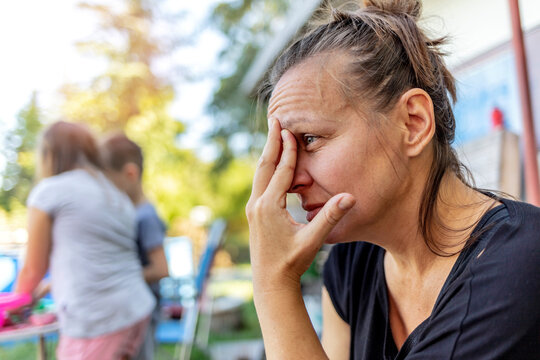Stressed depressed young mother sits outside in a playground with her kids playing behind her. Single frustrated woman hold her head with hands sitting on chair with playful kids on a background.