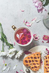 Heart shaped pastry with berries and pink flowers tea in white tea cup on white concrete kitchen table with flowers. Cozy, romantic tea time at home. Top view.