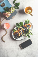 Feminine breakfast lifestyle with blueberries pancakes, cup of tea , burning candle and smartphone with black screen, blue handbag with hydrangea on light grey concrete table. Top view.