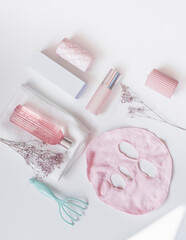 Modern facial skin care at home with sheet mask, facial toner, hydrating cream, candles and massage equipment on white background. Pink themed spa setting. Top view.