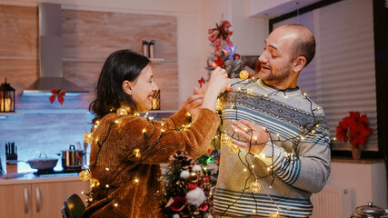 Festive couple trying to untangle garland of twinkle lights while decorating kitchen. Man and woman...