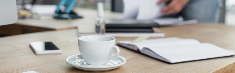 Cropped view of cup of coffee near cellphone, notebook and blurred businessman, banner