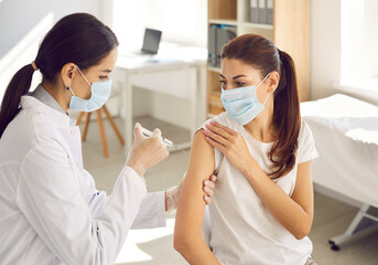 Female nurse in gloves giving a flu shot to a patient during a seasonal vaccination campaign. Beautiful young woman wearing a mouth covering getting a Covid-19 vaccine at a modern medical center - obrazy, fototapety, plakaty