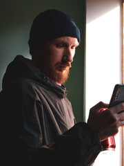 Bearded millennial man in black raincoat with smartphone in hand texting against wall with harsh shadow. Authentic male traveler Blogger guy lifestyle Solo travel adventure. Blogging communication