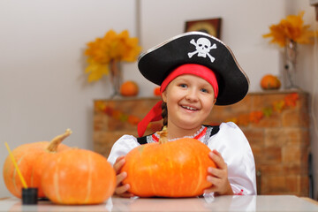 Halloween at home. A little girl dressed as a pirate sits at a table with pumpkins. The child smiles and looks into the camera. - obrazy, fototapety, plakaty