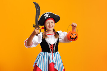 Halloween concept. Cheerful girl in a carnival costume of a pirate in the studio on a yellow background. The child is holding a saber and a bucket of pumpkin jack lantern.