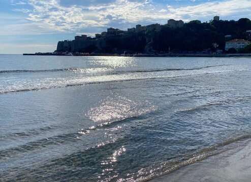 Sea waves on beach at evening sunset. Old town scenic cityscape. Beautiful silver shiny seascape. Mediterranean coast view. Relax, vacation, summer, happiness.