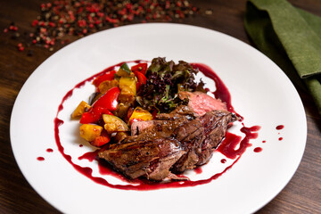 Chateaubriand of tender beef in Bordeaux wine sauce with vegetable sauté. Large plate of food on a dark brown wooden backdrop.