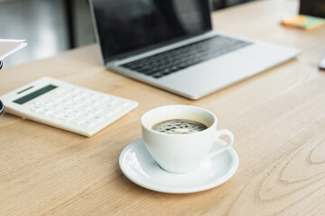 Cup of coffee near blurred laptop and calculator in office