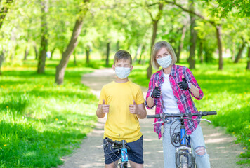 Mother and her young son wearing medical protective masks ride bikes in a summer park and show thumbs up gesture. Empty space for text