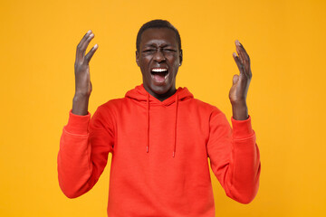 Frustrated irritated young african american man guy in red streetwear hoodie posing isolated on yellow background. People lifestyle concept. Mock up copy space. Screaming swearing, spreading hands.