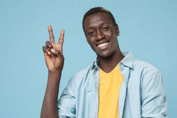 Fototapeta premium Cheerful young african american man guy in casual shirt, yellow t-shirt posing isolated on blue background studio portrait. People emotions lifestyle concept. Mock up copy space. Showing victory sign.
