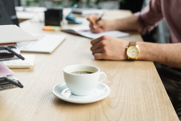 Cropped view of cup of coffee on table near blurred businessman in office