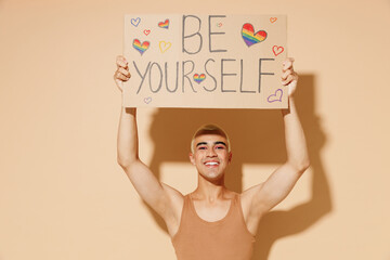 Young smiling happy cool blond latin gay man with make up in beige tank shirt hold card sign with...