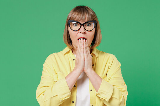 Elderly astonished surprised amazed shocked caucasian woman 50s wearing glasses yellow shirt cover mouth with hand isolated on plain green color background studio portrait People lifestyle concept