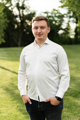 portrait of a young guy in a white shirt walking in the park at sunset. European guy looks at the camera. man on the background of the lawn in the park.