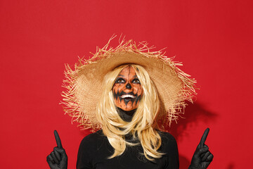 Young woman with Halloween makeup mask wears straw hat black scarecrow costume point fingers overhead on workspace isolated on plain red background studio portrait. Celebration holiday party concept.