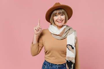 Traveler tourist fun mature elderly senior lady woman 55 years old wears brown shirt hat scarf hold index finger up with great new idea isolated on plain pastel light pink background studio portrait.