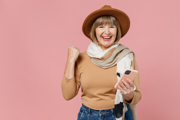 Traveler tourist mature elderly senior lady woman 55 years old wear brown shirt hat scarf hold use mobile cell phone clench fist say yes isolated on plain pastel light pink background studio portrait