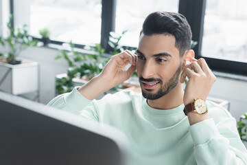 Smiling arabian businessman holding wireless earphone near blurred computer monitor in office