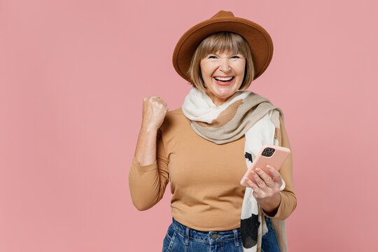 Traveler Tourist Mature Elderly Senior Lady Woman 55 Years Old Wear Brown Shirt Hat Scarf Hold Use Mobile Cell Phone Clench Fist Say Yes Isolated On Plain Pastel Light Pink Background Studio Portrait