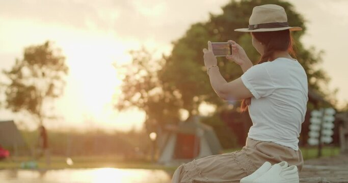 A female tourist in a brown hat, white T-shirt and brown skirt sits on a wooden bridge by the lake, using a mobile phone to take a beautiful view at sunset, an orange light reflecting on water surface