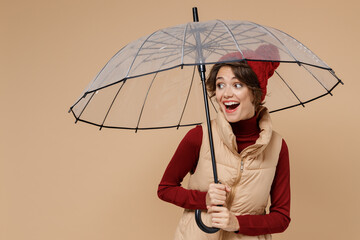 Young smiling surprised woman wears red turtleneck vest beret hold transparent umbrella look aside isolated on plain pastel beige background studio portrait. People lifestyle rainy weather concept - obrazy, fototapety, plakaty