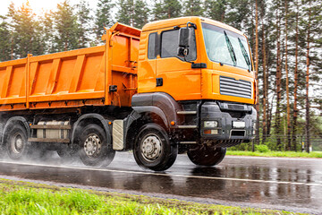 An orange 4-axle cargo dump truck with a load of 40 tons drives on a wet road in summer, in the...